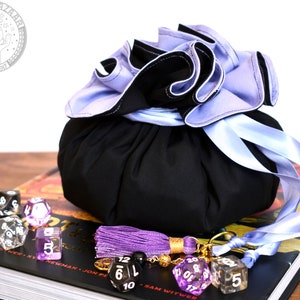 May include: A black dice bag with a purple and blue ruffled top and a matching ribbon. The bag is sitting on a book with a purple tassel and dice.