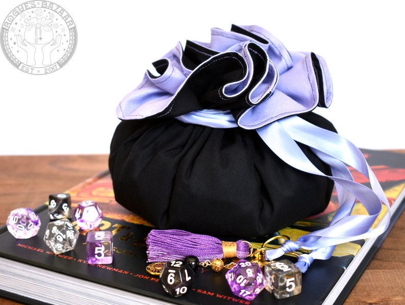 May include: A black dice bag with a purple and blue ruffled top and a matching ribbon. The bag is sitting on a book with a purple tassel and dice.