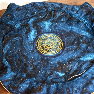 May include: A round, padded mat with a celestial design. The fabric features a swirling pattern of blue and black, speckled with gold. A gold embroidered emblem is centered on the mat, which has a cream-colored border.