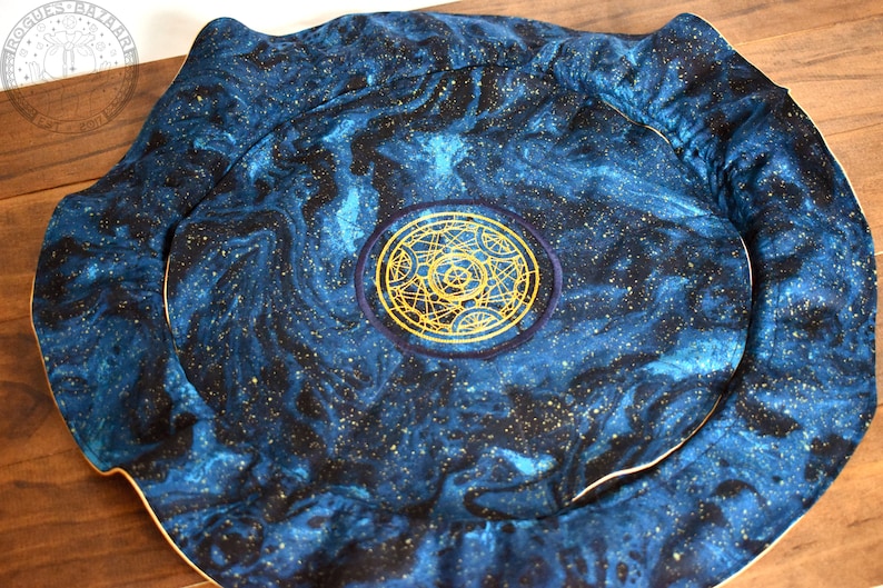May include: A round, padded mat with a celestial design. The fabric features a swirling pattern of blue and black, speckled with gold. A gold embroidered emblem is centered on the mat, which has a cream-colored border.