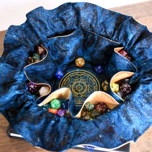May include: A blue fabric dice bag with a celestial pattern, filled with various polyhedral dice. The bag has multiple pockets and a gold-colored embroidered design in the center. The bag is open, displaying the dice.