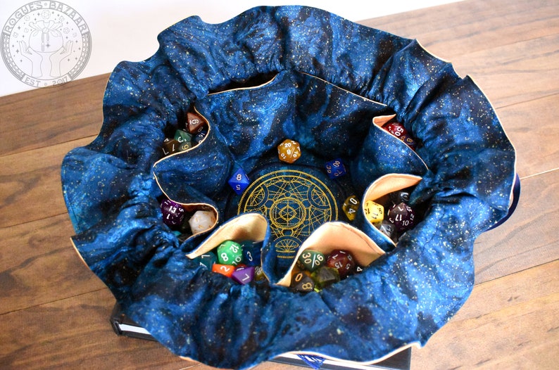 May include: A blue fabric dice bag with a celestial pattern, filled with various polyhedral dice. The bag has multiple pockets and a gold-colored embroidered design in the center. The bag is open, displaying the dice.