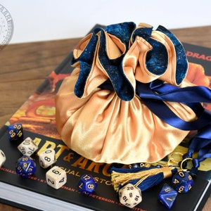 May include: A gold satin dice bag with a navy blue velvet trim and drawstring, resting on a book titled "Art & Arcana". Several dice in various colors and styles are scattered around the bag, with a navy blue tassel.