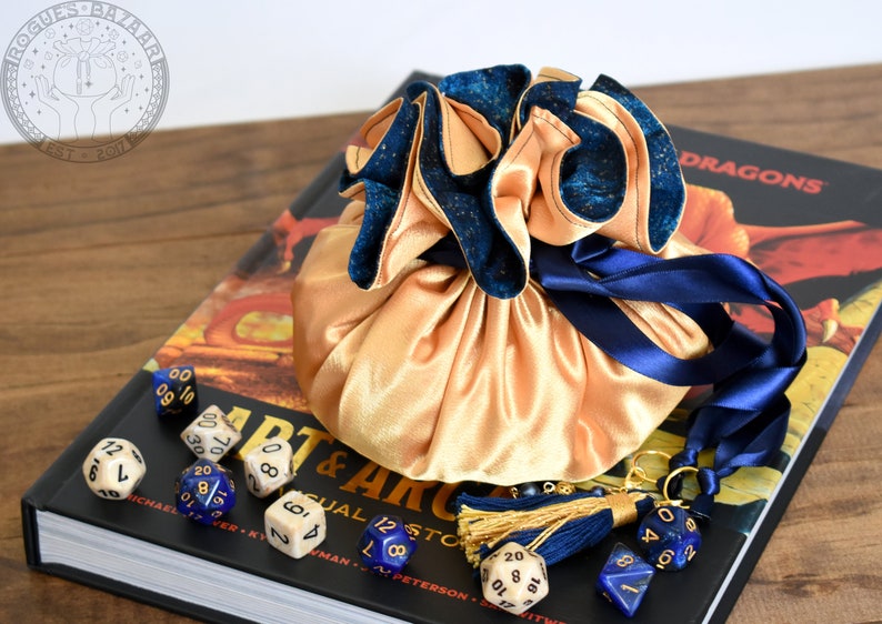 May include: A gold satin dice bag with a navy blue velvet trim and drawstring, resting on a book titled "Art & Arcana". Several dice in various colors and styles are scattered around the bag, with a navy blue tassel.