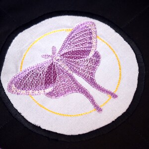 May include: A purple embroidered moth with a yellow circle around it on a white background. The moth is on a black fabric.