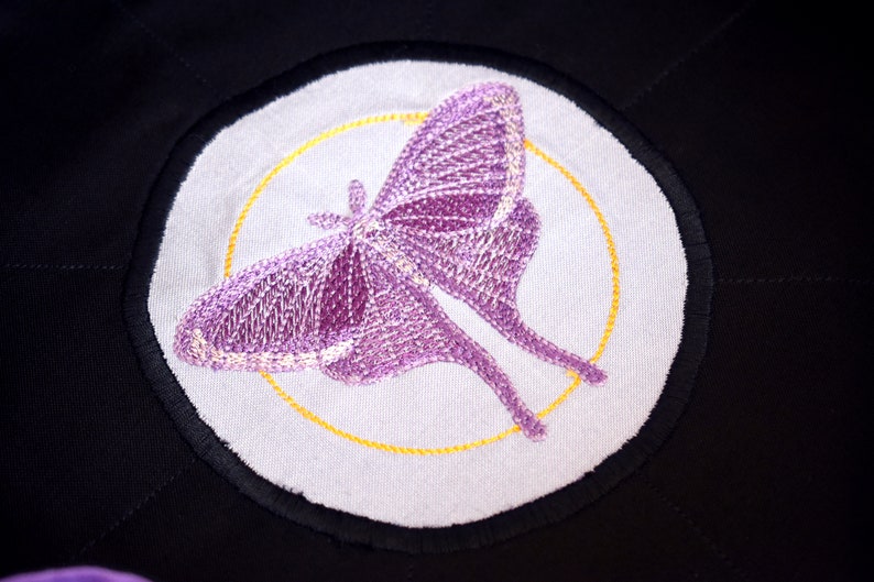 May include: A purple embroidered moth with a yellow circle around it on a white background. The moth is on a black fabric.