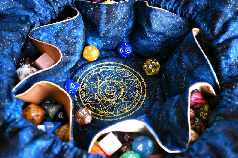 May include: A blue dice bag with a celestial pattern, filled with various multi-sided dice in different colors. The bag has a gold embroidered design in the center. The dice include numbers like 3, 6, 12, and 20.