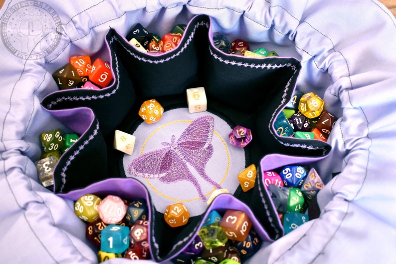 May include: A purple and black dice bag with a moth design. The bag has six compartments for dice and a central compartment for a larger die. The bag is filled with a variety of colorful dice.