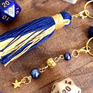 May include: A navy blue and gold tassel keychain with gold-colored hardware. The keychain features a star charm, dark blue beads, and a decorative gold ball. The tassel is made of navy blue and gold threads.