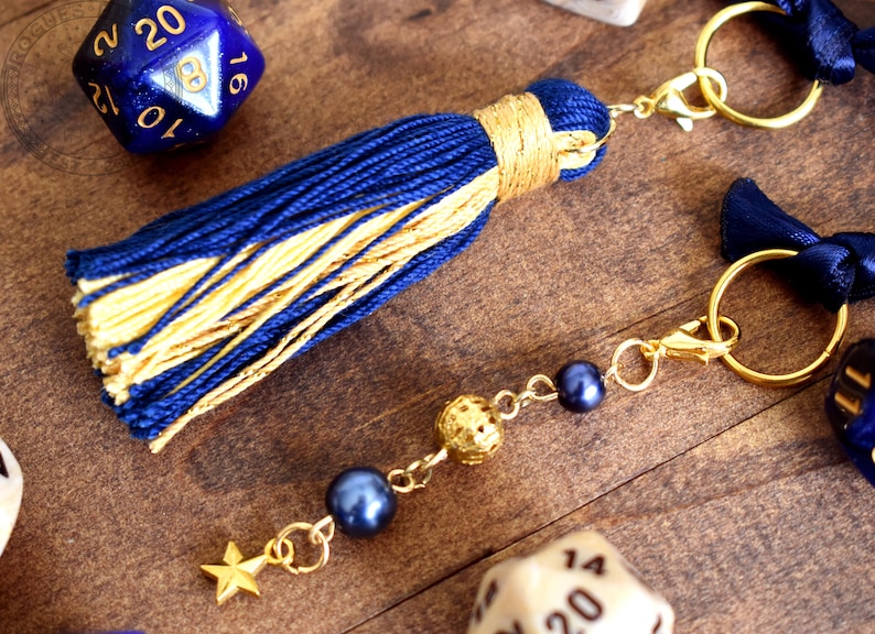 May include: A navy blue and gold tassel keychain with gold-colored hardware. The keychain features a star charm, dark blue beads, and a decorative gold ball. The tassel is made of navy blue and gold threads.