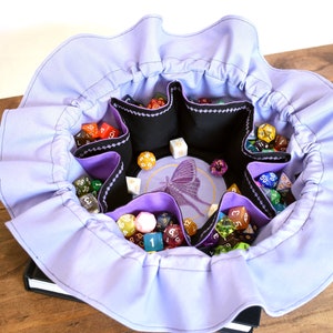 May include: A lavender fabric dice bag with a ruffled edge. The bag has six compartments for dice and a central embroidered design of a butterfly. The bag is filled with colorful dice.