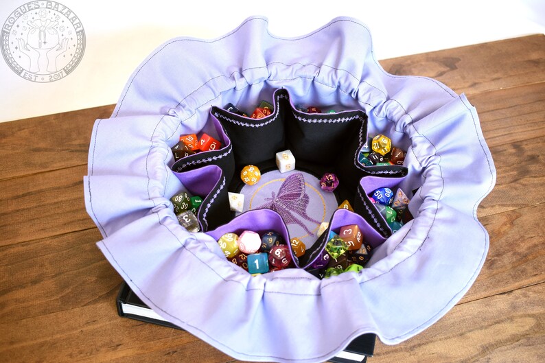May include: A lavender fabric dice bag with a ruffled edge. The bag has six compartments for dice and a central embroidered design of a butterfly. The bag is filled with colorful dice.