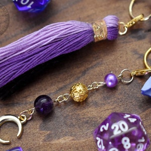 May include: Purple and black tassel keychain with a gold crescent moon charm, amethyst bead, gold bead, and a purple pearl. The keychain is attached to a silver chain and a gold ring.
