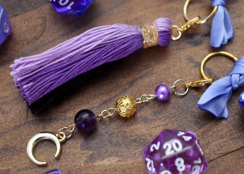 May include: Purple and black tassel keychain with a gold crescent moon charm, amethyst bead, gold bead, and a purple pearl. The keychain is attached to a silver chain and a gold ring.