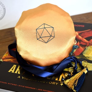 May include: A golden-colored dice bag with a blue embroidered geometric design, resting on a book titled "ARKHAM: A VISUAL HISTORY". A navy blue ribbon and gold tassel with charms are attached to the bag. The bag is on a wooden surface.