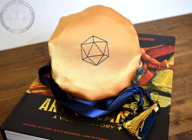 May include: A golden-colored dice bag with a blue embroidered geometric design, resting on a book titled "ARKHAM: A VISUAL HISTORY". A navy blue ribbon and gold tassel with charms are attached to the bag. The bag is on a wooden surface.