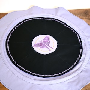 May include: A round, lavender fabric cushion with a black center circle. The center circle has a purple butterfly embroidered on it. The cushion is perfect for meditation or yoga.