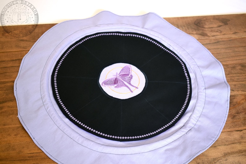 May include: A round, lavender fabric cushion with a black center circle. The center circle has a purple butterfly embroidered on it. The cushion is perfect for meditation or yoga.
