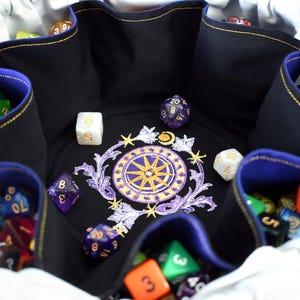 May include: A black dice bag with a blue interior and gold trim, filled with colourful polyhedral dice. The bag features an embroidered design of a compass rose with floral accents. The bag is open, displaying the dice.