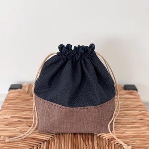 May include: A drawstring bag with a dark blue top and a brown bottom. The bag has a small, repeating pattern of white circles on the blue fabric. The bag is sitting on a woven wicker surface.