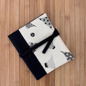 May include: A rectangular, cream-colored fabric case with a black border and tie closure. The case features a repeating black geometric pattern. The case is resting on a wooden surface.