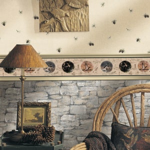 Op de afbeelding: Een beige behang met een bruine houtsnijwerk van vogels en een rand met een herhalend patroon van vogels en dennenappels. Het behang wordt geaccentueerd door een muur met een stenen look, een houten stoel en een bijzettafel met een lamp en dennenappels.