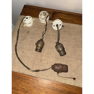 May include: A collection of vintage light fixtures, including three white porcelain sockets and three brass bulb holders. The fixtures are connected by aged wiring, with one fixture featuring a pull chain. The items are arranged on a tan surface.