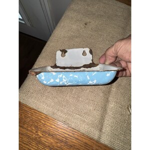 May include: Vintage blue and white enamelware soap dish with a speckled pattern. The dish has a rectangular shape with a raised back and a rusty metal bracket for wall mounting. The dish is held by a hand.