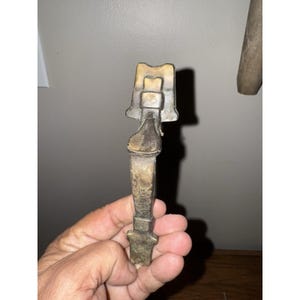 May include: A vintage metal bottle opener with a unique design. The opener has a long handle and a decorative top. The metal has a tarnished, aged appearance, suggesting it is an antique or vintage item.