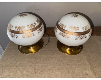 Może przedstawiać: Dwie lampy kuliste w stylu vintage z białymi szklanymi kloszami. Każdy klosz ma złoty pas z kwiatowymi wzorami i złotą podstawę. Lampy stoją na beżowej powierzchni.