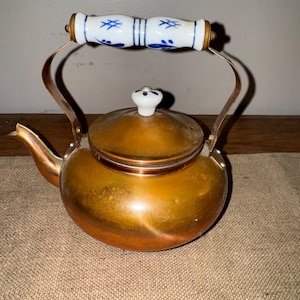 May include: A copper teapot with a white and blue patterned handle and lid. The teapot has a rounded body and a curved spout. The handle is made of ceramic and metal. The teapot is likely vintage and suitable for kitchen decor.