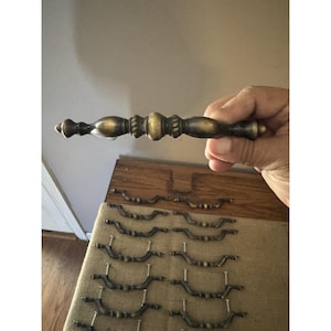 May include: A collection of antique brass drawer pulls and handles. The image shows a variety of styles, including a larger, ornate handle held in a hand, and several smaller, curved pulls with screws attached. The hardware has a vintage, aged appearance.