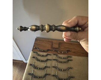 May include: A selection of antique brass drawer pulls and handles. The image displays various styles, including a larger, ornate handle held in a hand, and several smaller, curved pulls with screws attached. The hardware has a vintage, aged appearance.