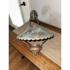 May include: A wall-mounted lamp featuring a weathered, verdigris-toned metal shade with a scalloped edge. The fixture includes a central burner component with a warm, glowing light source and a circular ring pull at the bottom, suspended by metal arms.