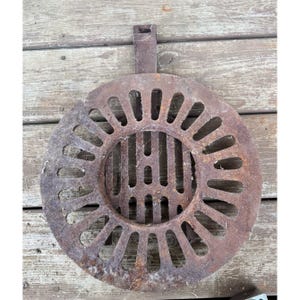 May include: A rusty, circular metal grate with a handle. The grate features a series of radial slots around a central, slotted area. The metal has a weathered appearance, suggesting outdoor use and age. The handle is attached to the top.