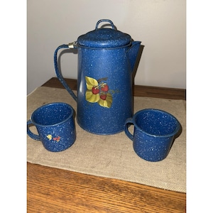 May include: A blue speckled enamelware coffee pot with a lid, handle, and spout, accompanied by two matching mugs. The coffee pot features a strawberry design. This vintage set is perfect for camping or rustic decor.