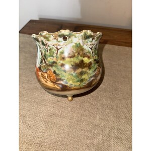 May include: A small, round, cream-colored ceramic planter with a scalloped rim and a single hole for hanging. The exterior features a hand-painted landscape scene with green trees, brown foliage, and gold-painted feet at the base of the vessel.