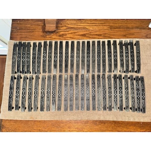 May include: A collection of fifty-two rectangular metal drawer pulls arranged in two rows. Each pull features a dark, weathered finish with a repeating embossed geometric pattern consisting of interconnected diamond and floral shapes along the center.