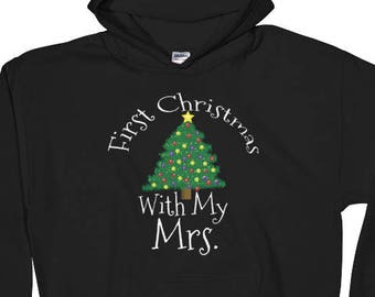 May include: Black hooded sweatshirt with the text "First Christmas With My Mrs." above a decorated Christmas tree graphic. The tree is green with yellow and red ornaments and a yellow star on top. A festive holiday apparel item.