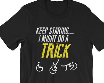 May include: Black t-shirt with the text "KEEP STARING... I MIGHT DO A TRICK" in yellow. The text is in a distressed font. There are three white stick figure icons below the text, one in a wheelchair, one doing a backflip, and one doing a cartwheel.