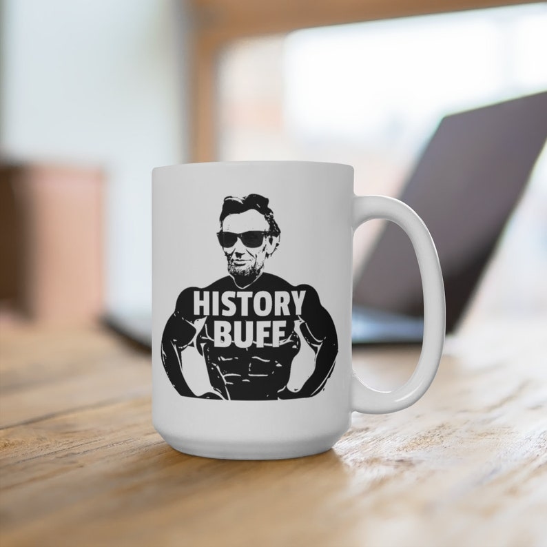 May include: White ceramic coffee mug with a black and white illustration of Abraham Lincoln with sunglasses and the text "HISTORY BUFF".