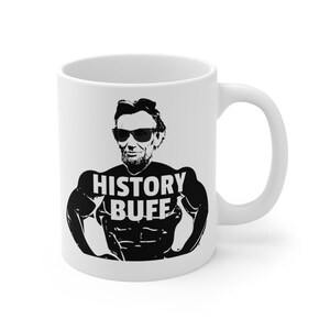 May include: White ceramic coffee mug with a black and white image of Abraham Lincoln wearing sunglasses and a muscle-bound physique. The text "HISTORY BUFF" is printed on the mug.