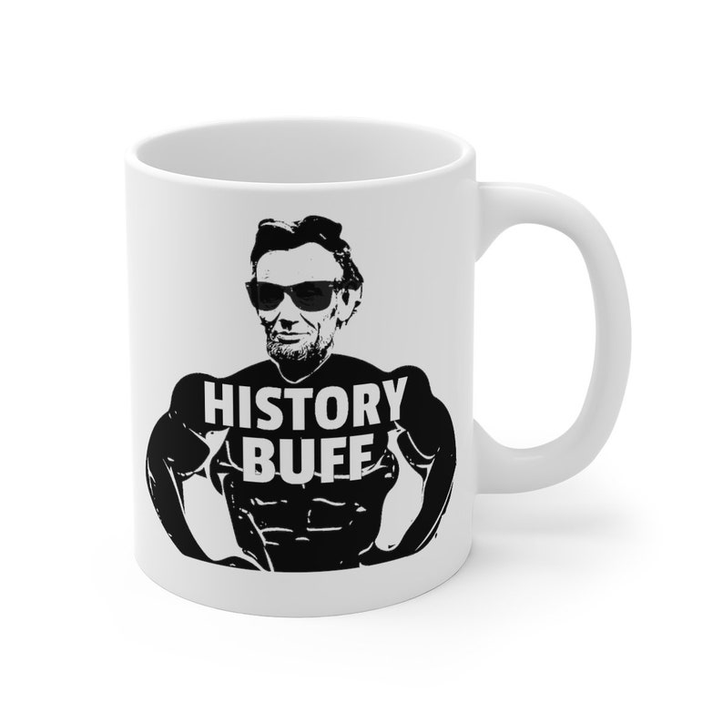 May include: White ceramic coffee mug with a black and white image of Abraham Lincoln wearing sunglasses and a muscle-bound physique. The text "HISTORY BUFF" is printed on the mug.