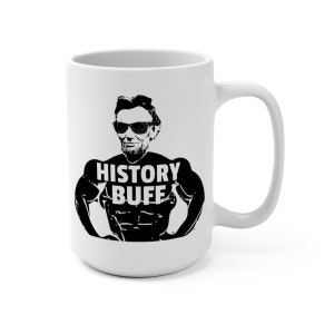 May include: White ceramic coffee mug with a black and white illustration of Abraham Lincoln wearing sunglasses and a muscle shirt with the text "HISTORY BUFF".