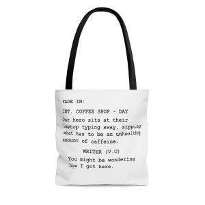 May include: White tote bag with black handles featuring a script-style text design that reads "FADE IN: INT. COFFEE SHOP - DAY Our hero sits at their laptop typing away, sipping what has to be an unhealthy amount of caffeine. WRITER (V.O) You might be wondering how I got here."
