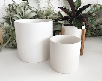 May include: Two white ceramic plant pots with wooden stands. The larger pot is taller and wider than the smaller pot.