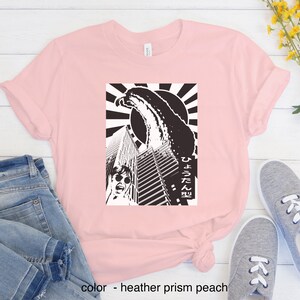May include: A pink t-shirt featuring a black and white graphic print of a monster attacking a city. The graphic includes the Japanese text "ひょろろん" which translates to "slender".