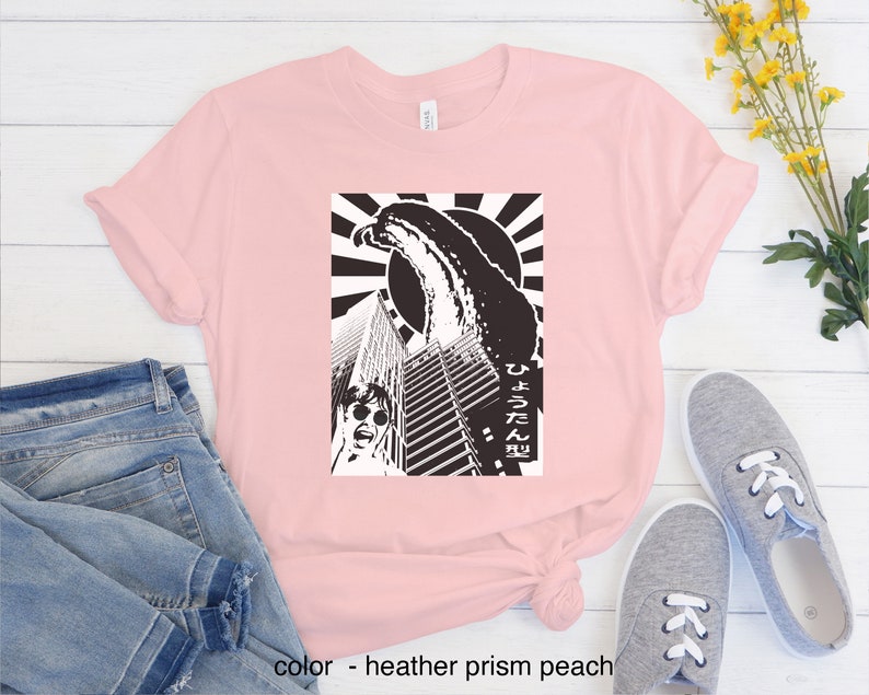 May include: A pink t-shirt featuring a black and white graphic print of a monster attacking a city. The graphic includes the Japanese text "ひょろろん" which translates to "slender".
