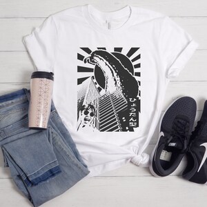 May include: White t-shirt featuring a black and white graphic print of a monster attacking a city. The graphic includes Japanese text that reads "ひょろたん型".
