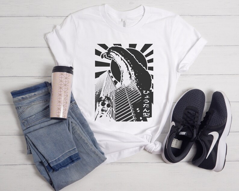May include: White t-shirt featuring a black and white graphic print of a monster attacking a city. The graphic includes Japanese text that reads "ひょろたん型".